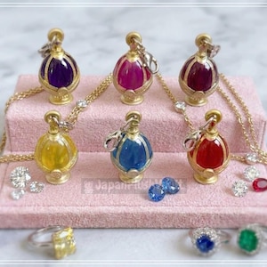 Könnte beinhalten: Eine Sammlung goldfarbener Anhänger-Halsketten mit bunten, mit Edelsteinen gefüllten Flaschenanhängern. Die Anhänger werden auf einer rosa Oberfläche präsentiert, mit Diamant- und Edelsteinringen und losen Edelsteinen.