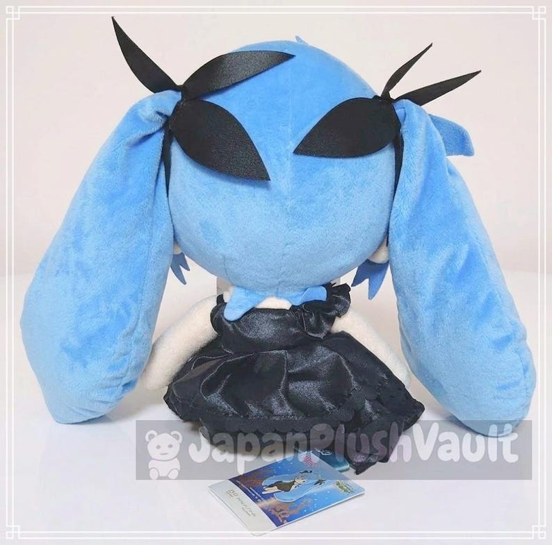 May include: A plush doll with long, light blue hair and black accents. The doll has a black dress and is holding a small card. The doll's hair is styled with black ribbons. The doll is a soft, cuddly toy.