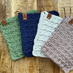 May include: Four handmade crochet items in shades of green, navy blue, light grey, and mauve. Each item has a leather tag with a gold button and a leather loop for hanging. The items are arranged on a wooden surface.