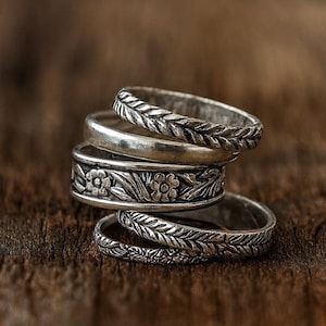 May include: Five silver rings with detailed patterns are stacked on a wooden surface. The rings showcase floral and leaf motifs, with a braided texture on some. The rustic wooden background enhances the vintage appeal of the jewellery.