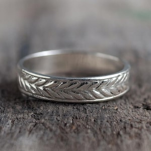 Könnte beinhalten: Ein silberner Ring mit einem detaillierten Blattmuster um das Band. Der Ring ist auf einer rustikalen Holzoberfläche platziert, was sein aufwendiges Design und seinen metallischen Glanz hervorhebt. Das Design des Rings ist klassisch.