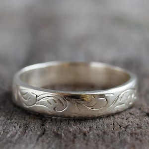 Könnte beinhalten: Ein silberner Ring mit einem eingravierten floralen Muster. Der Ring ist auf einer rustikalen Holzoberfläche platziert. Das Band hat eine glatte, polierte Oberfläche, und das florale Design ist um den gesamten Umfang geätzt. Der Ring ist ein Schmuckstück.