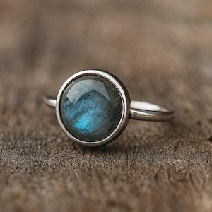 Può includere: Un anello d'argento con una gemma rotonda blu iridescente. La pietra è incastonata in una semplice montatura a castone, e il gambo è sottile e lucido. L'anello è esposto su una superficie di legno rustico, evidenziando il contrasto.