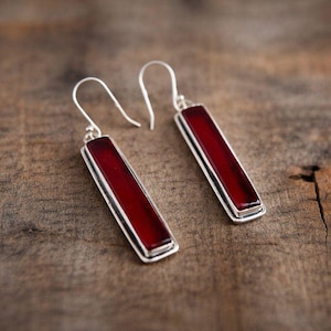 Puede incluir: Un par de pendientes rectangulares con un centro rojo oscuro y un borde plateado. Los pendientes cuelgan de un gancho plateado. Los pendientes se exhiben sobre una superficie de madera.
