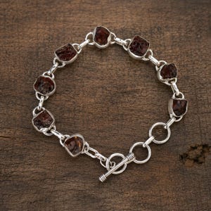 May include: A silver chain bracelet featuring eight rough, dark red garnet stones set in individual metal frames. The bracelet is secured with a toggle clasp consisting of a circular ring and a T-shaped bar, connected by several small silver links.