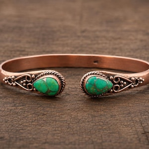 May include: A copper cuff bracelet featuring two teardrop-shaped green turquoise stones at the open ends. The metal band is decorated with intricate wire scrollwork and twisted rope detailing surrounding each stone setting.