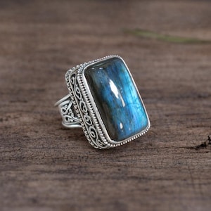 May include: A silver ring featuring a large, rectangular labradorite gemstone with a blue and teal iridescence. The ring has an ornate, filigree design around the stone and on the band.