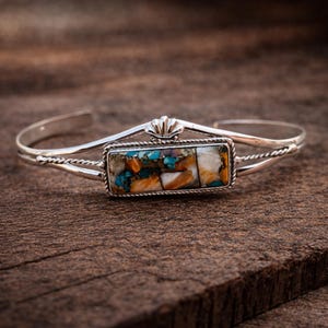 May include: A silver cuff bracelet with a rectangular turquoise and orange stone centerpiece. The bracelet has a decorative shell-like design at the top and a twisted rope pattern on the sides. The bracelet is displayed on a wooden surface.