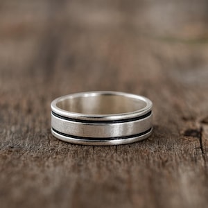 Könnte beinhalten: Ein silberner Ring mit zwei dünnen, schwarzen Bändern. Der Ring ist vor einem verschwommenen Holzgrund platziert. Das Design des Rings ist schlicht und modern, geeignet für den täglichen Gebrauch.