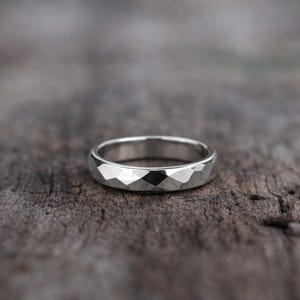 Könnte beinhalten: Ein silberner Ring mit facettiertem Design. Der Ring hat ein geometrisches Muster aus kleinen, abgewinkelten Oberflächen, die das Licht reflektieren. Der Ring ist vor einem verschwommenen Holzgrund platziert.