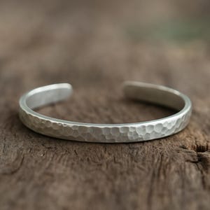 Op de afbeelding: Een zilveren manchetarmband met een gehamerde textuur. Het open ontwerp heeft een reeks kleine, gelijkmatig verdeelde inkepingen over het oppervlak. De armband wordt op een houten oppervlak gepresenteerd.