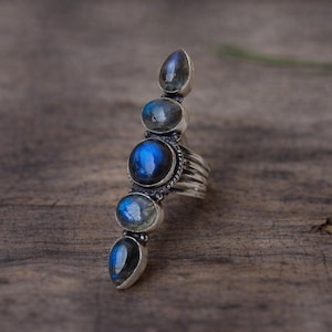 Puede incluir: Un anillo de plata con cinco piedras preciosas de labradorita ovaladas alineadas verticalmente. Las piedras muestran destellos iridiscentes azules y grises. La banda de metal tiene un acabado texturizado y oxidado con un detalle de cuerda.