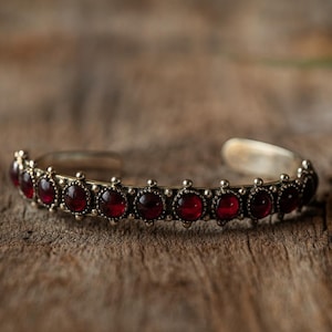 May include: A silver-toned cuff bracelet with a row of deep red, round gemstones. The bracelet has a detailed, ornate design with small, decorative accents between each gemstone. The background is a textured, wooden surface.