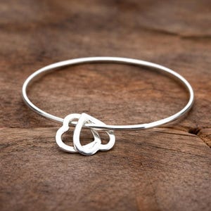 Könnte beinhalten: Ein silbernes Armreif-Armband mit zwei verbundenen Herzanhängern. Das Armband besteht aus dünnem, glänzendem Metall und liegt auf einer Holzoberfläche. Die Herzen sind glatt und poliert und verleihen dem Design einen Hauch von Eleganz.