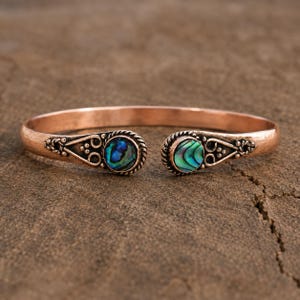 Könnte beinhalten: Ein Kupfer-Armreif im offenen Design mit zwei runden Abalone-Muschel-Einlagen an den Enden. Das Metallband weist eine texturierte, dunkle Oberfläche mit detaillierten seilartigen Verzierungen und Schnörkeln um jede schillernde Muschel herum auf.