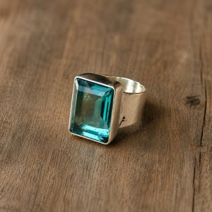 Pode incluir: Um anel de prata com uma grande pedra preciosa azul de corte esmeralda retangular, engastada numa moldura de metal lisa. A banda é larga e polida, apresentando um acabamento metálico limpo que complementa a pedra azul vibrante e translúcida.