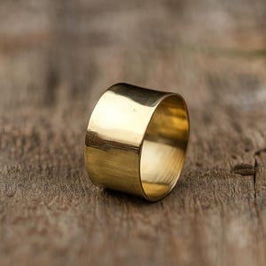 May include: A wide, gold-colored ring with a smooth, polished surface. The ring is a solid band, reflecting light and set against a rustic wooden background. The ring's design is simple and elegant.