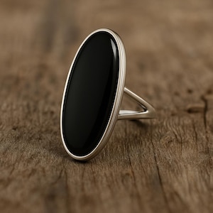 May include: A silver ring featuring a large, oval black stone. The stone is set in a polished silver band, creating a striking contrast. The ring's design is simple yet elegant, with a focus on the bold black gemstone. The ring is displayed on a wooden surface.