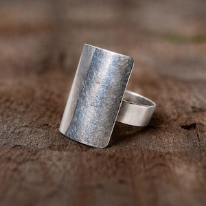 May include: A silver-colored ring with a rectangular face. The ring has a brushed metal texture and a wide band. The ring is set against a brown wooden surface.