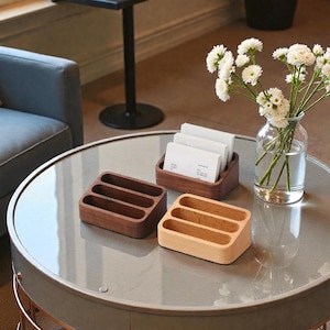 May include: Three wooden business card holders in varying shades of brown and natural wood are displayed on a round table. One holder is filled with white business cards. A vase of white flowers sits nearby.