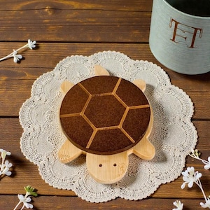 May include: A wooden turtle-shaped coaster with a brown, geometric shell design and light-colored bamboo body. The coaster sits on a white lace doily, with a light blue pot in the background. Decorative and functional.