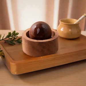 May include: A wooden tray holds a dark brown sphere nestled in a matching wooden holder. A small yellow ceramic pot with a straw sits nearby. The scene is bathed in warm, natural light, suggesting a focus on natural materials and home decor.