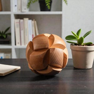 May include: A wooden puzzle ball with a complex, interlocking design. The ball is made of light brown wood and has a spherical shape with multiple curved and angular pieces. The puzzle sits on a dark surface, with a small potted plant in the background.