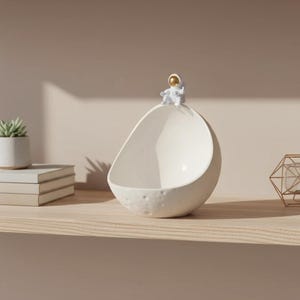 May include: A white ceramic bowl shaped like a crescent moon, with a textured surface. A small astronaut figurine with a gold helmet sits on top. The bowl is on a wooden shelf, with a succulent plant and books in the background.
