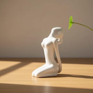Könnte beinhalten: Eine weiße Keramikfigur, die eine kniende Figur mit minimalistischem Design darstellt. Die Skulptur hat glatte Kurven und eine glänzende Oberfläche. Die Figur kniet auf einer Holzoberfläche, mit einem grünen Blatt im Hintergrund.