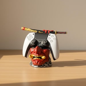 Może przedstawiać: Biało-czarny kontroler do gier wideo spoczywa na czerwono-złotej podstawie maski demona. Miniaturowa czarno-złota katana umieszczona jest na górze kontrolera. Kontroler i podstawa znajdują się na drewnianej powierzchni.
