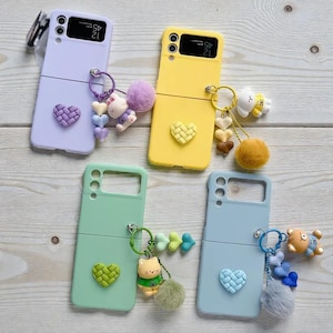 May include: Four brightly coloured phone cases, each with a heart-shaped design and a keychain. The cases are in shades of lilac, yellow, mint green, and pale blue. Each case has a different keychain with a small plush toy and a pom-pom.