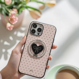 May include: A pink phone case with a black polka dot pattern and a black heart-shaped phone grip. The phone case has a black border and a silver ring around the heart. The word "Bonnie" is printed on the bottom of the case.
