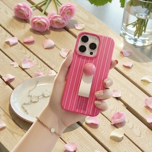 May include: A pink phone case with vertical white stripes and a matching pink pop socket. The case is held in a hand, with a bracelet and a silver necklace visible in the background. Pink flower petals and flowers are scattered around.