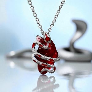 May include: A silver-toned necklace featuring a pendant with a red heart-shaped stone wrapped by a silver snake. The chain is a delicate link style. The background is a light blue gradient with a blurred silver cobra.