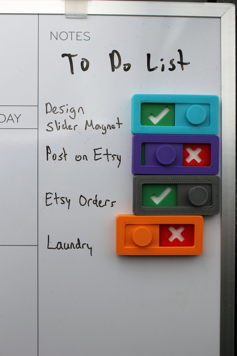 May include: A white board with a handwritten "To Do List" and a list of tasks with corresponding toggle switches. The tasks are: Design Slider Magnet, Post on Etsy, Etsy Orders, and Laundry. The toggle switches are color-coded and have checkmarks or X marks to indicate completion status.