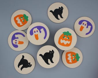 May include: A collection of round Halloween-themed coasters. The coasters feature illustrations of a black cat, a jack-o'-lantern, and a ghost in orange, purple, and green on a cream background. The coasters are arranged on a light gray surface.