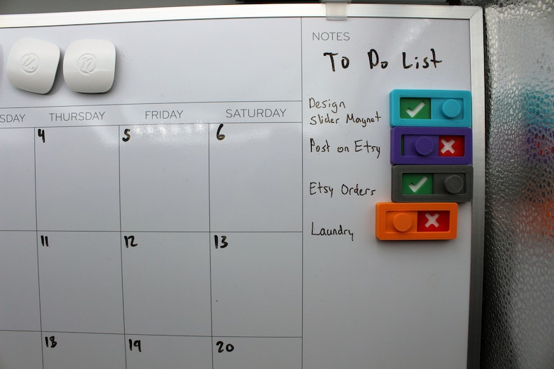 May include: A white dry-erase board with a calendar grid and a to-do list. The to-do list includes "Design Slider Magnet", "Post on Etsy", "Etsy Orders", and "Laundry". Each task has a colored square with a checkmark or an X to indicate completion status.