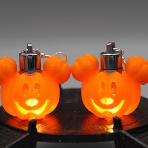May include: Two orange Mickey Mouse-shaped Halloween lights with silver tops and ear details. Each light has a smiling face and is illuminated. The lights are on a black surface, perfect for Halloween decor.