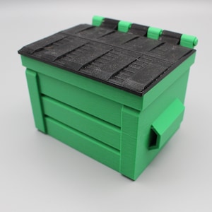 May include: A green plastic toy dumpster with a black lid. The lid has a textured surface and is hinged on the top.