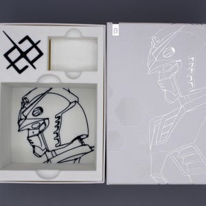 May include: A white box with a black and white illustration of a robot's head and a matching design on the box cover. The box is open, revealing the robot head illustration inside. The box has a gray background.