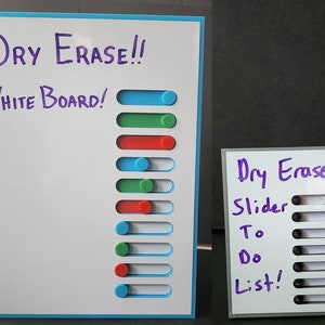 May include: Two white dry erase boards with blue and gray frames. The larger board has the text "DRY ERASE!! WHITE BOARD!" written in purple marker. The smaller board has the text "Dry Erase! Slider To Do List!" written in purple marker. Both boards have rows of white slider bars with colored circles that can be moved along the bars.