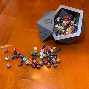 May include: A gray, 20-sided die with numbers 1-20 printed on each side. The die is open and filled with a variety of colorful dice. The die is sitting on a wooden table.