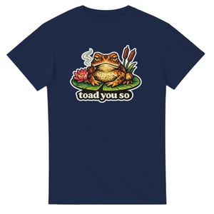 May include: Navy blue t-shirt with a cartoon frog smoking a pipe, sitting on a lily pad and the text "toad you so". The frog is brown and yellow with an angry expression. A pink flower and cattails are also on the lily pad.