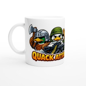 May include: White ceramic mug featuring a cartoon design of two ducks in military gear, one with an axe and the other with a machine gun. The mug has the text "QUACK ATTACK" in orange and yellow.