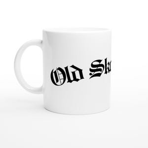 May include: White ceramic mug featuring the words "Old Skool" in a black gothic font. The mug has a curved handle and a shiny finish. The design is centered on the mug.
