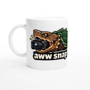 以下が含まれることがあります： 白いセラミックマグカップ。漫画のワニガメのデザイン。茶色と緑色のワニガメがカメラを持っています。下には「aww snap」の文字。写真愛好家向けの面白いアイテムです。