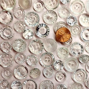 May include: A collection of clear glass buttons in various shapes and sizes. The buttons are arranged in a random pattern on a light pink surface.