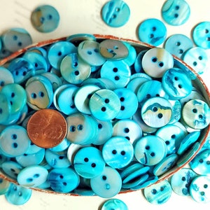 May include: A bowl filled with turquoise blue and white shell buttons. The buttons have two holes and are arranged in a random pattern. There is a penny in the bowl for scale.