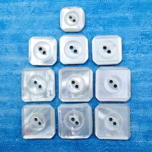 May include: A collection of ten square, white mother-of-pearl buttons arranged in a pyramid formation. Each button features a smooth, iridescent surface with a central circular indentation and two small holes for threading, showcasing natural luster.