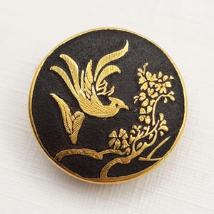 May include: Round black button with a gold bird and floral design. The bird is perched on a branch with flowers. The button has a gold border.